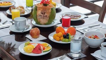 A breakfast table set with plates of fruit, a coconut, and beverages, with a view of greenery in the background.