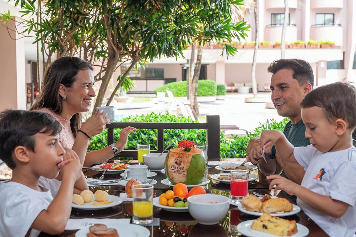 Una familia desayunando al aire libre en una mesa con frutas, pasteles y una bebida de coco.