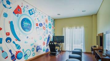A room with a wall mural featuring various icons, a gaming chair, a large TV, and a gaming console.
