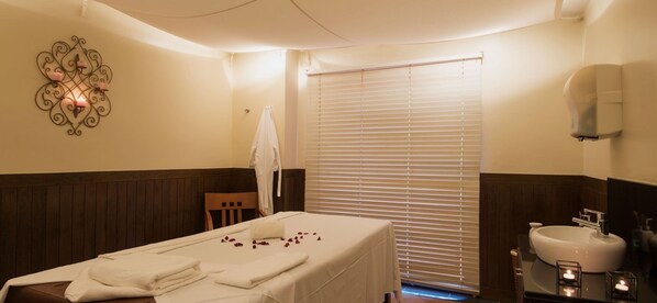 A spa room with a massage table, towels, a wall-mounted decorative piece, a window with blinds, and a sink with a soap dispenser. - Vila Gale Fortaleza (Fortaleza)