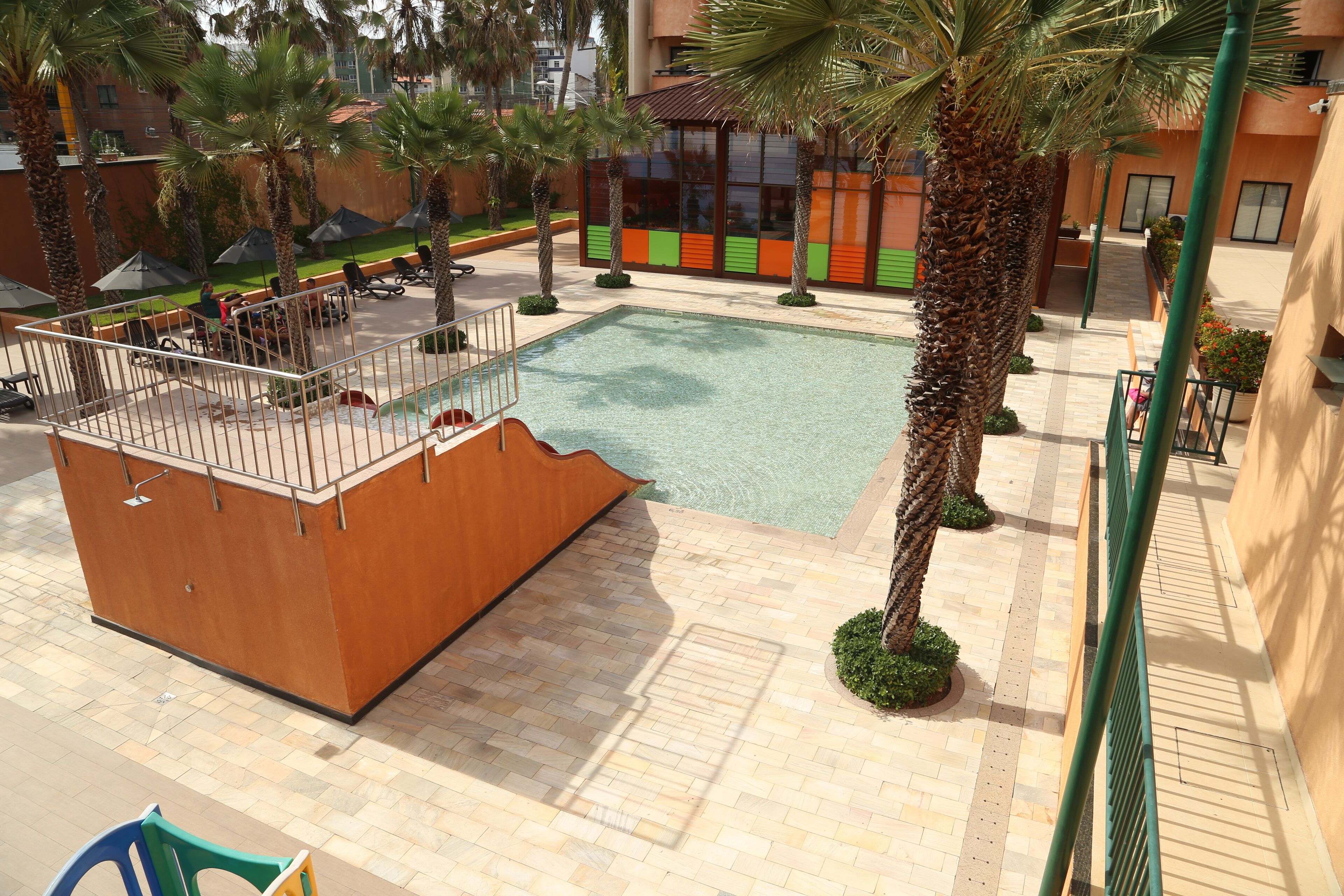 A pool area with a wooden deck, palm trees, and a building with colorful accents in the background.