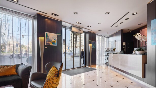 A modern hotel lobby with a reception desk, a black leather sofa, a patterned armchair, and a large window offering a view of the street. - Eurostars Monumental (Barcelona)