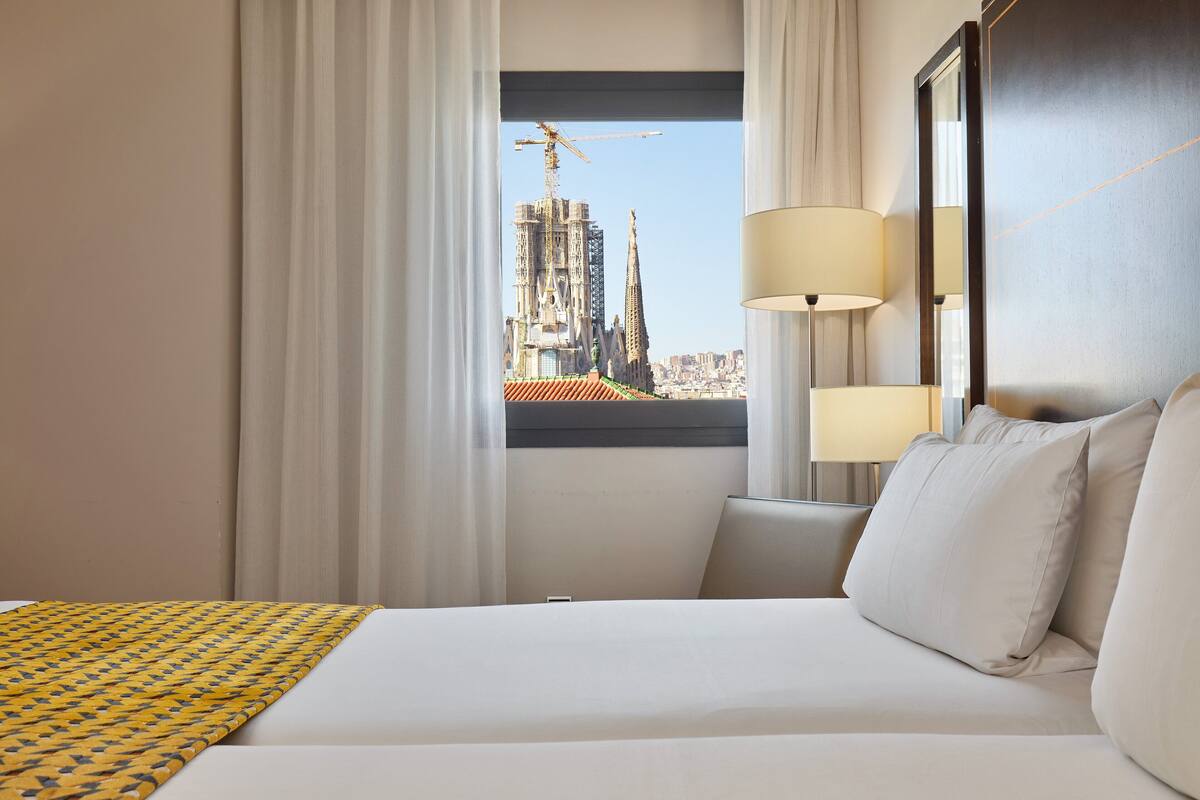 Une chambre d’hôtel avec un grand lit, deux lampes de chevet, une vue sur un paysage urbain avec un immeuble imposant, et une fenêtre avec des rideaux.