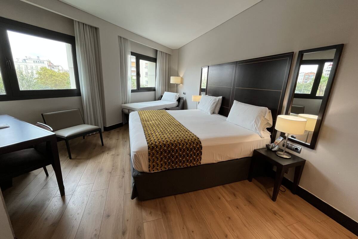 Une chambre d’hôtel avec un grand lit, un bureau et une chaise. Il y a deux fenêtres avec des rideaux, une table de chevet avec une lampe et un miroir au mur.