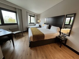 A hotel room with a large bed, a desk, and a chair. There are two windows with curtains, a bedside table with a lamp, and a mirror on the wall.