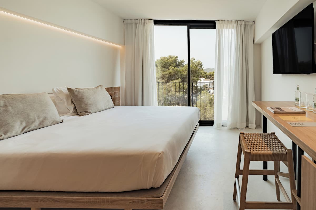 A hotel room with a large bed, a desk with a chair, a TV mounted on the wall, and a balcony with curtains.