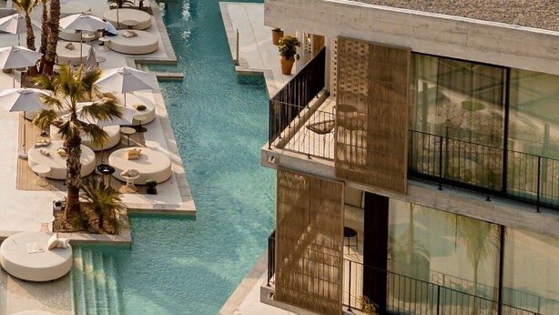 Un área de piscina en la azotea con sillones de descanso y sombrillas, una piscina azul cristalina y un edificio moderno con grandes ventanales.