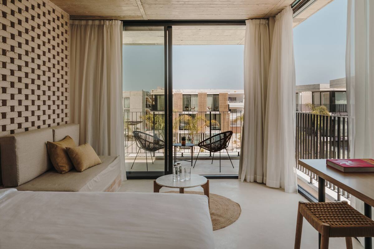A modern hotel room with a large bed, a sofa, a small round table, and a balcony with chairs.