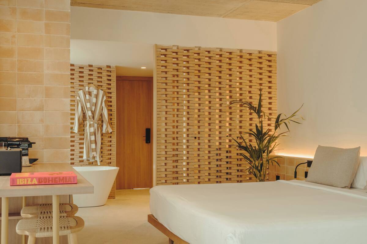 A modern hotel room with a wooden bed, a desk with a chair, a bathroom with a bathtub, and a large plant.
