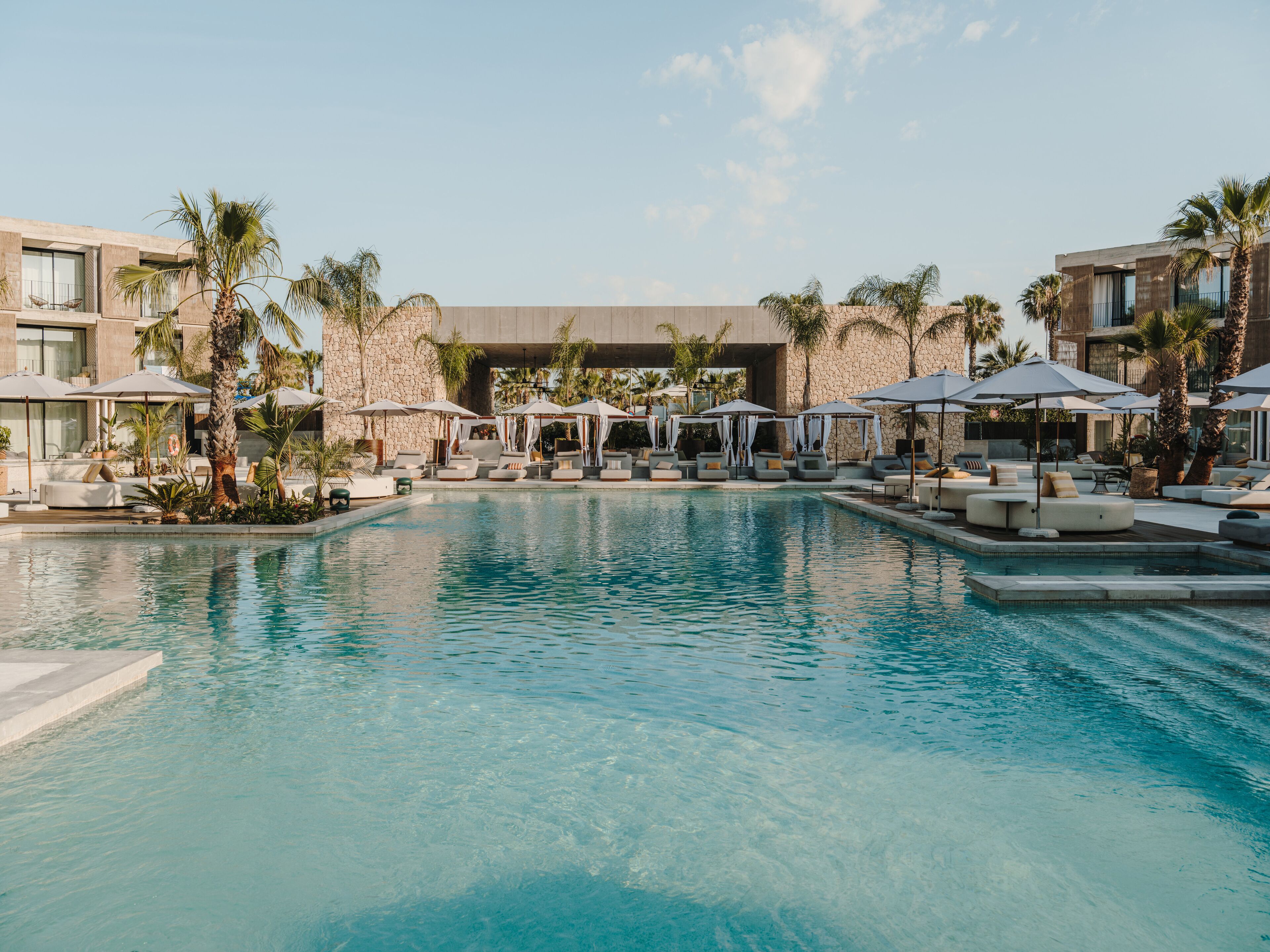 A swimming pool with lounge chairs and umbrellas, surrounded by palm trees and modern buildings.