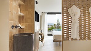Un baño moderno con un lavabo de piedra grande, un taburete de madera y vistas a un balcón con una cama y una bata blanca colgada en un perchero de madera.