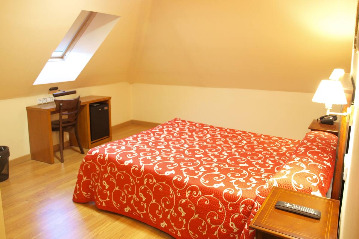 A bedroom with a bed, a desk, a chair, a small table with a lamp, and a ceiling light.