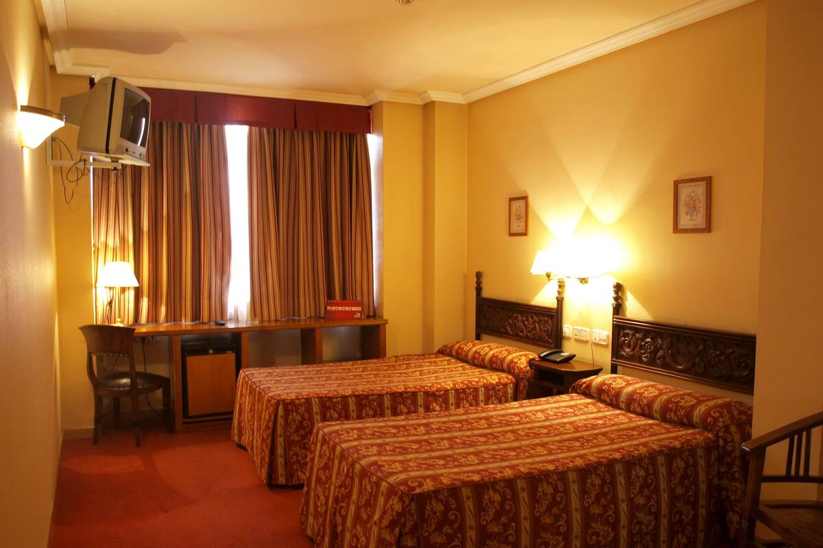 A hotel room with two beds, a desk, a chair, a TV, and a lamp.