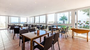 Een ruime, overdekte eethoek met meerdere tafels en stoelen, grote ramen met uitzicht op de omgeving, en een betegelde vloer.