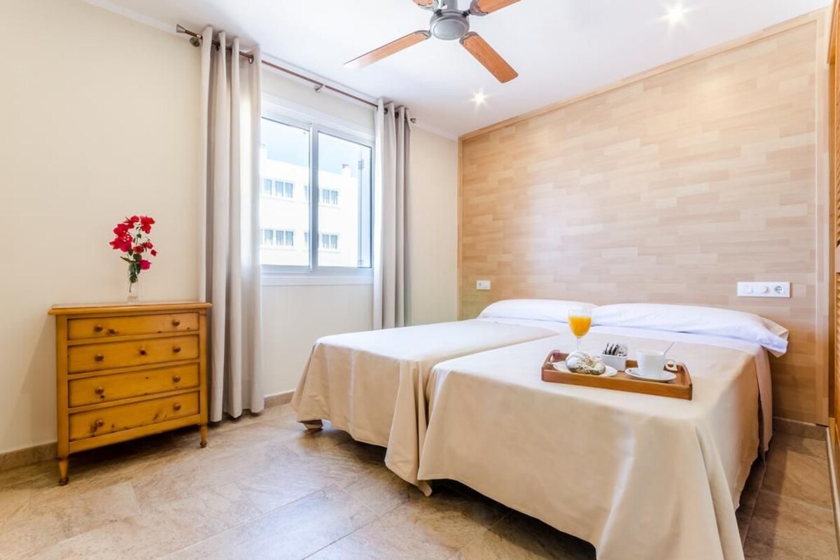 Habitación de hotel con dos camas, un aparador de madera, una ventana con cortinas, un ventilador de techo y una bandeja de desayuno sobre las camas.
