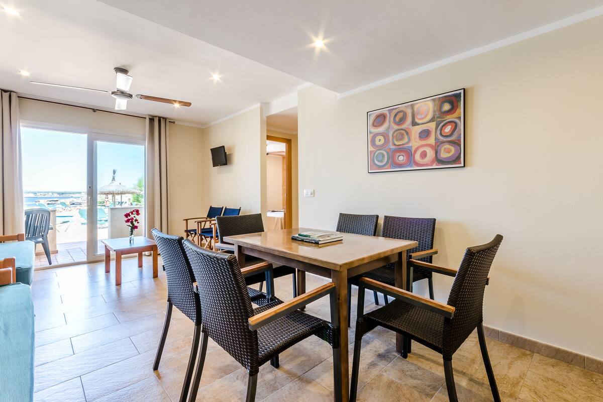 Un comedor moderno con una mesa de madera y sillas de mimbre, un televisor de pantalla plana en la pared y vistas al exterior a través de amplias ventanas.