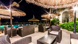 Een terras op het dak met rieten meubels, rieten parasols en een zwembad op de achtergrond.