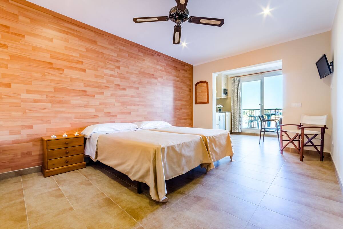 Un dormitorio con pared de madera, una cama con sábanas blancas, una mesita de noche con velas y un ventilador de techo.