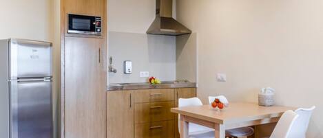 A compact kitchen with a refrigerator, microwave, and stove. There is a wooden cabinet with a microwave on top, a stainless steel range hood, and a small table with chairs.