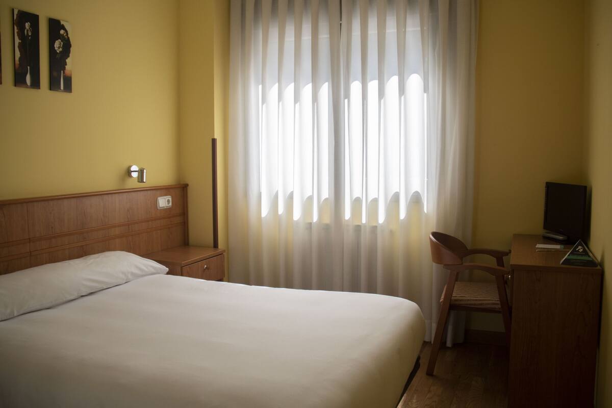 Une chambre d’hôtel comprenant un lit, un bureau avec une chaise, une télévision et une fenêtre avec des rideaux.