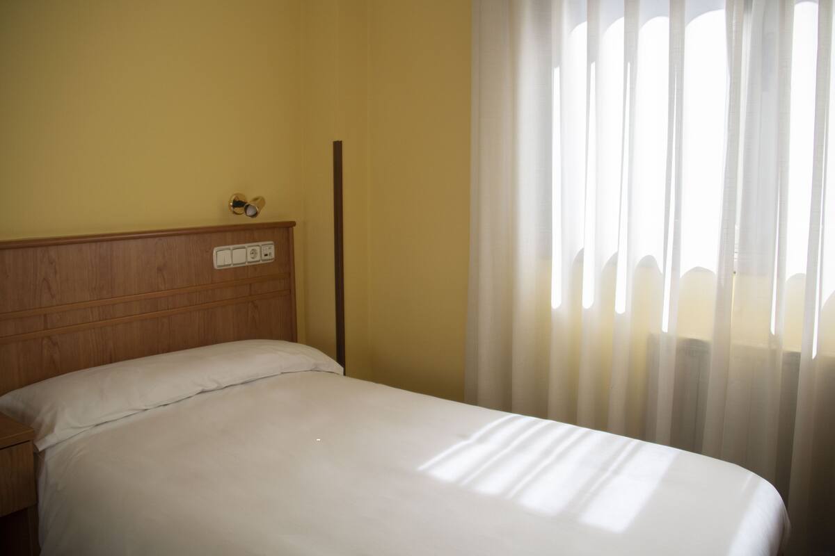 Une chambre d’hôtel avec une tête de lit en bois, un lit avec une literie blanche, une table de chevet et une fenêtre ornée de rideaux légers.