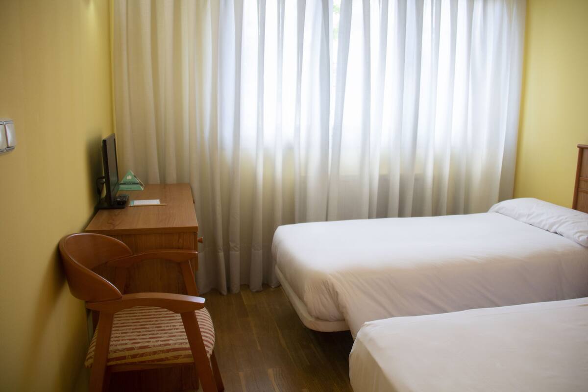 Une chambre d’hôtel avec deux lits simples, un bureau en bois avec une chaise et une fenêtre recouverte d’un rideau.