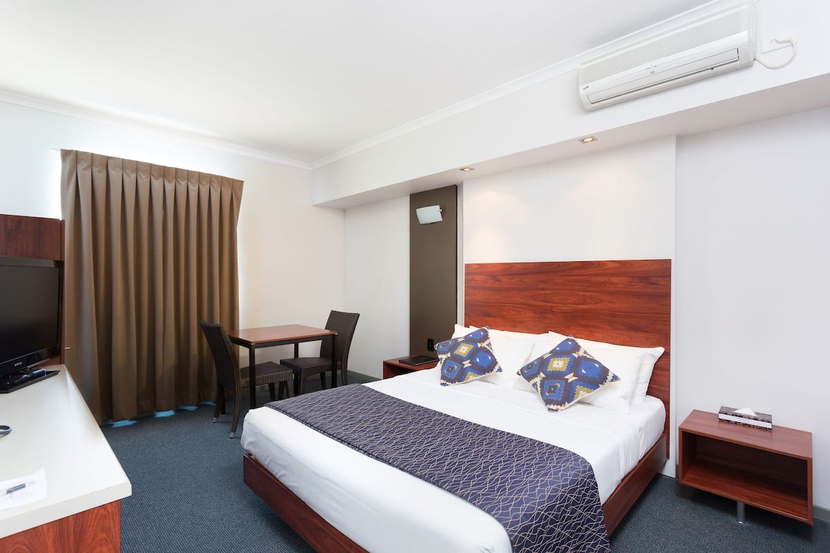 A hotel room with a bed, a desk, a chair, a TV, and an air conditioning unit.