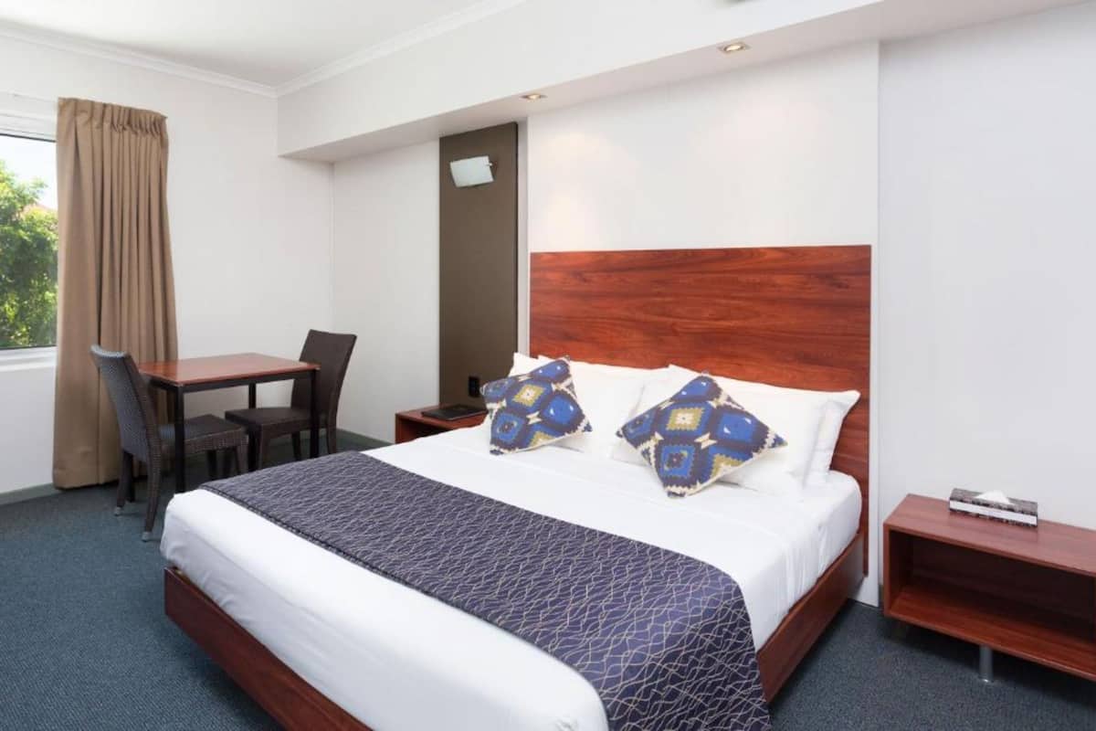 A hotel room with a large bed, a wooden headboard, two bedside tables, a desk with a chair, and a window with curtains.