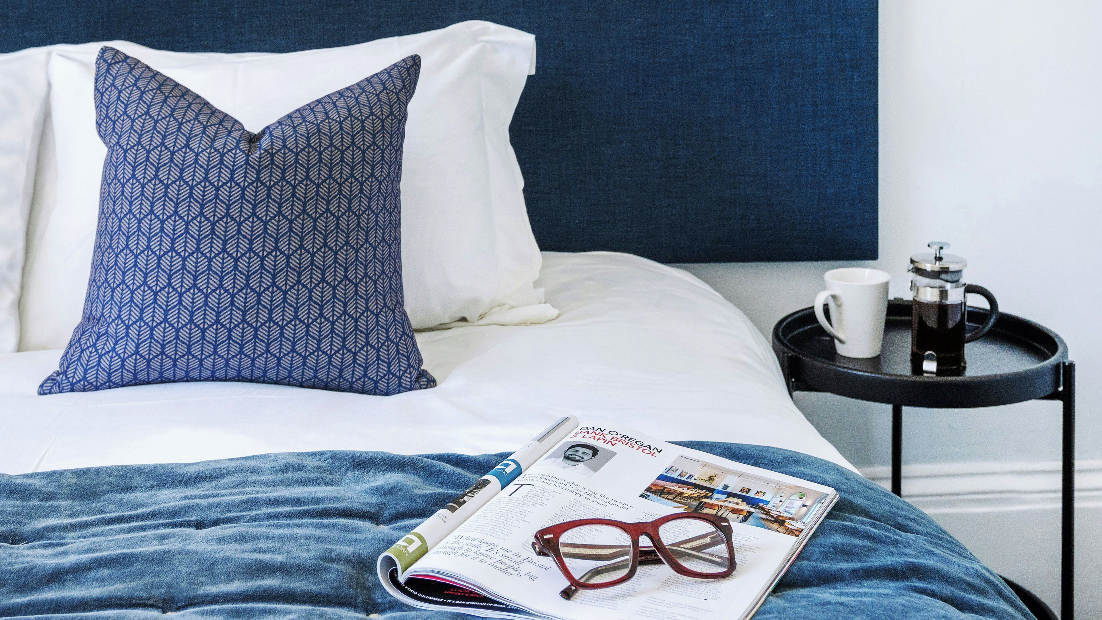 A neatly made bed with a patterned pillow, a magazine, and a pair of glasses on it.