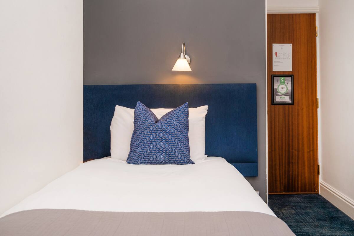 A neatly made bed with a blue headboard, a wall-mounted light fixture, and a door with a framed certificate.