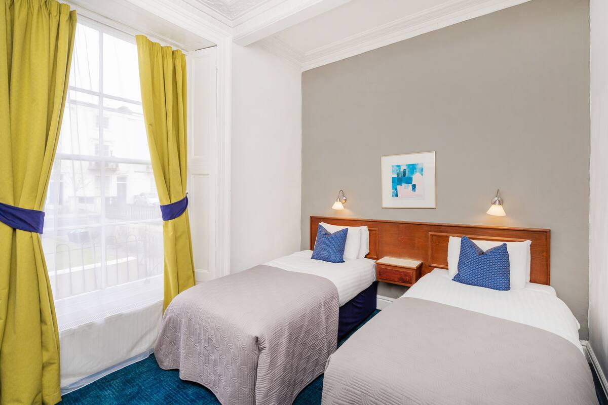 A hotel room with two single beds, a wooden headboard, a nightstand, a framed artwork on the wall, and a window with yellow curtains.