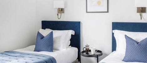 Habitación de hotel con dos camas, cada una con cabecero azul y ropa de cama blanca, una mesita redonda con cafetera y taza, y un cuadro en la pared.