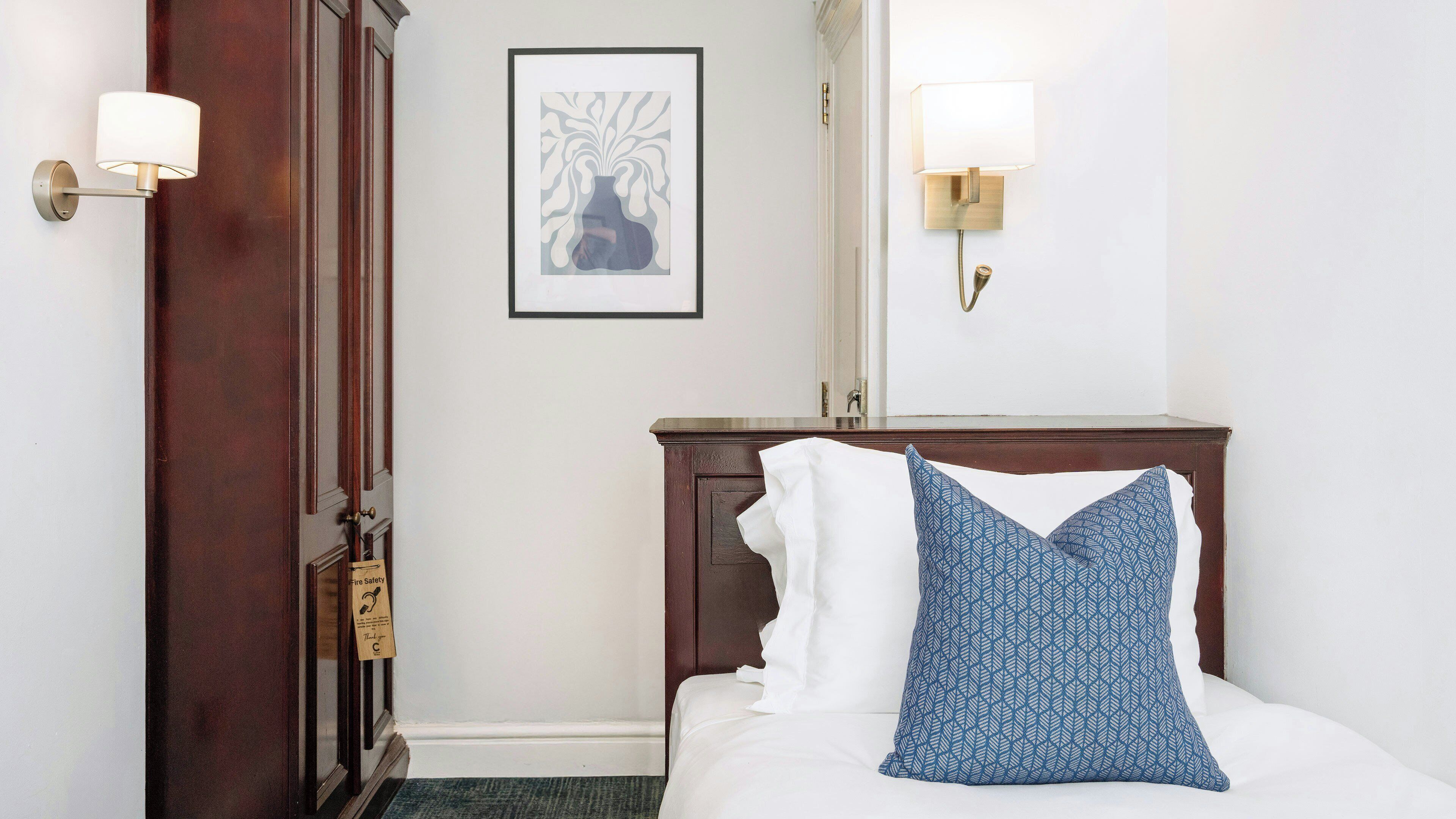 A hotel room with a wooden headboard, a framed artwork on the wall, a bedside table with a lamp, and a door with a safety sign.