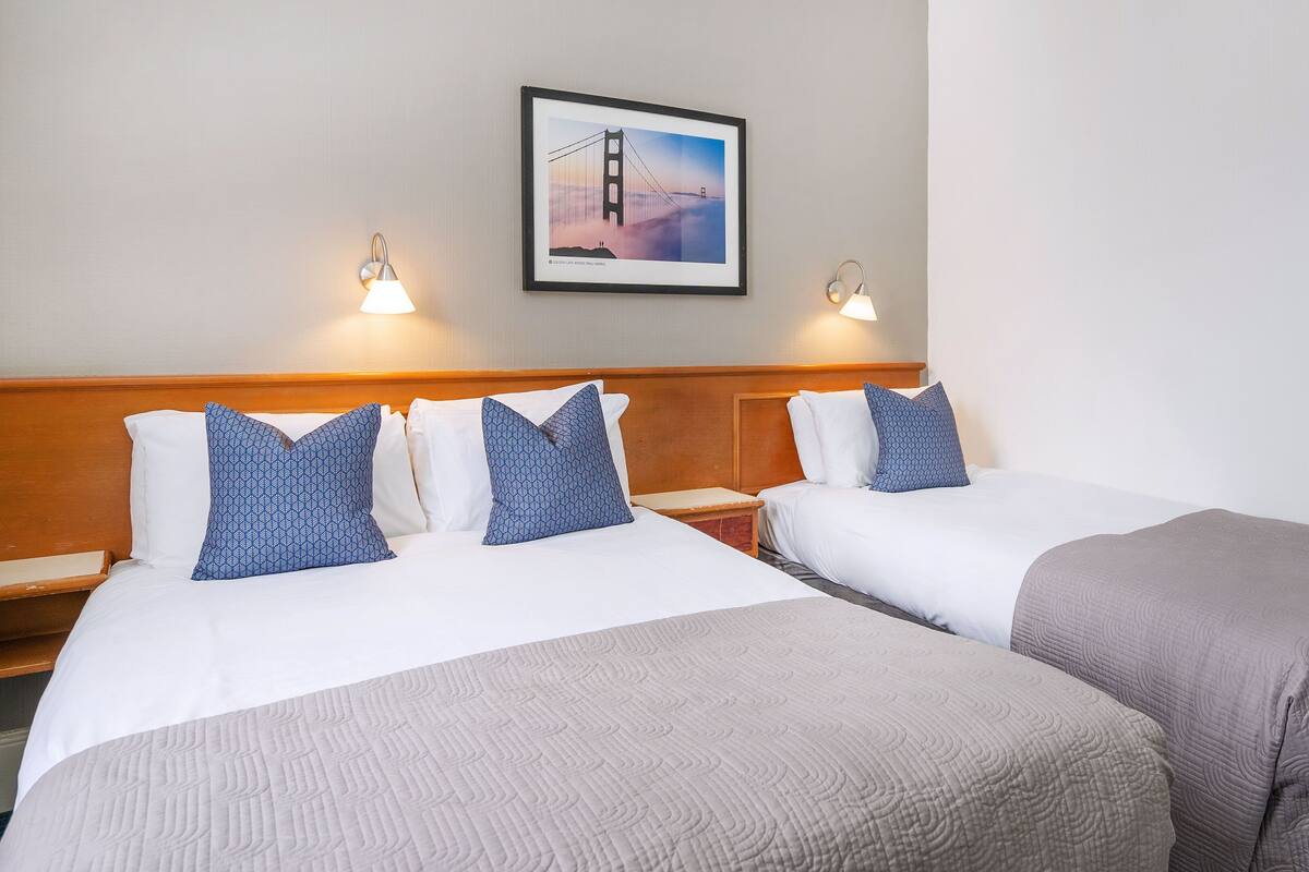 A hotel room with two beds, a wooden headboard, a framed picture of a bridge, and two wall-mounted lamps.