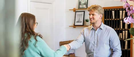 Una recepción con una persona de camisa a rayas estrechando la mano a otra. Se ve un reloj en la pared, un cuadro enmarcado y una estantería con libros y una planta en maceta al fondo.