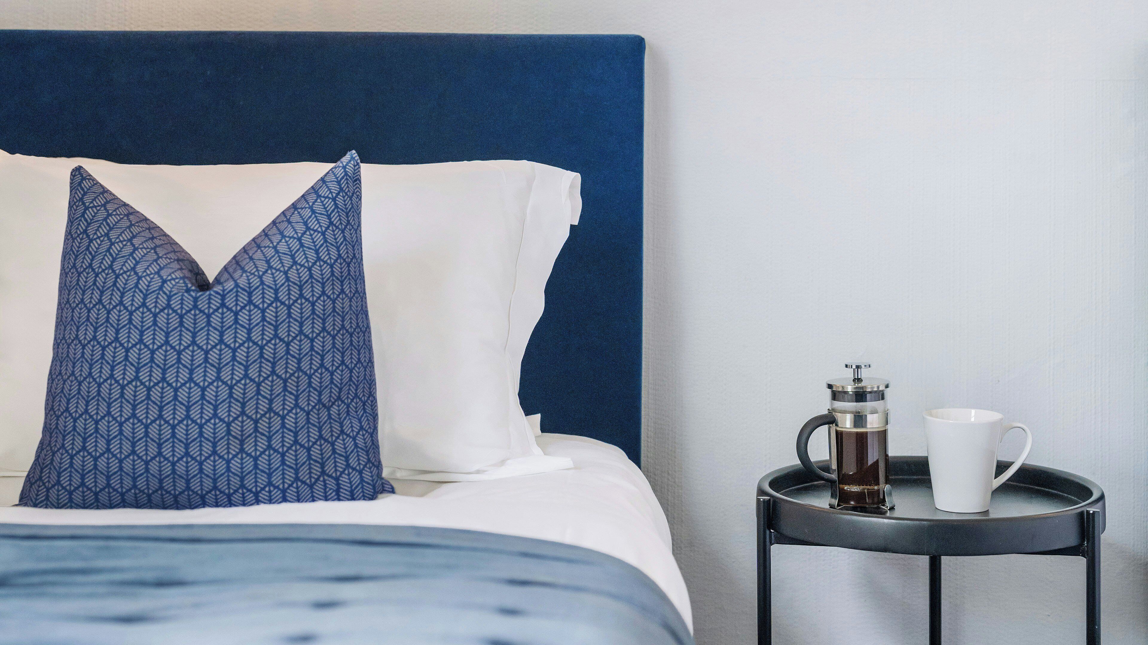 Una cama bien hecha con un cabecero azul, una almohada estampada y una almohada blanca. Junto a la cama hay una mesita con una cafetera y una taza blanca.