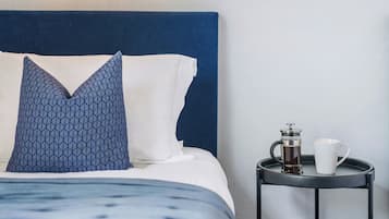 A neatly made bed with a blue headboard, a patterned pillow, and a white pillow. Beside the bed is a small table with a coffee maker and a white mug.