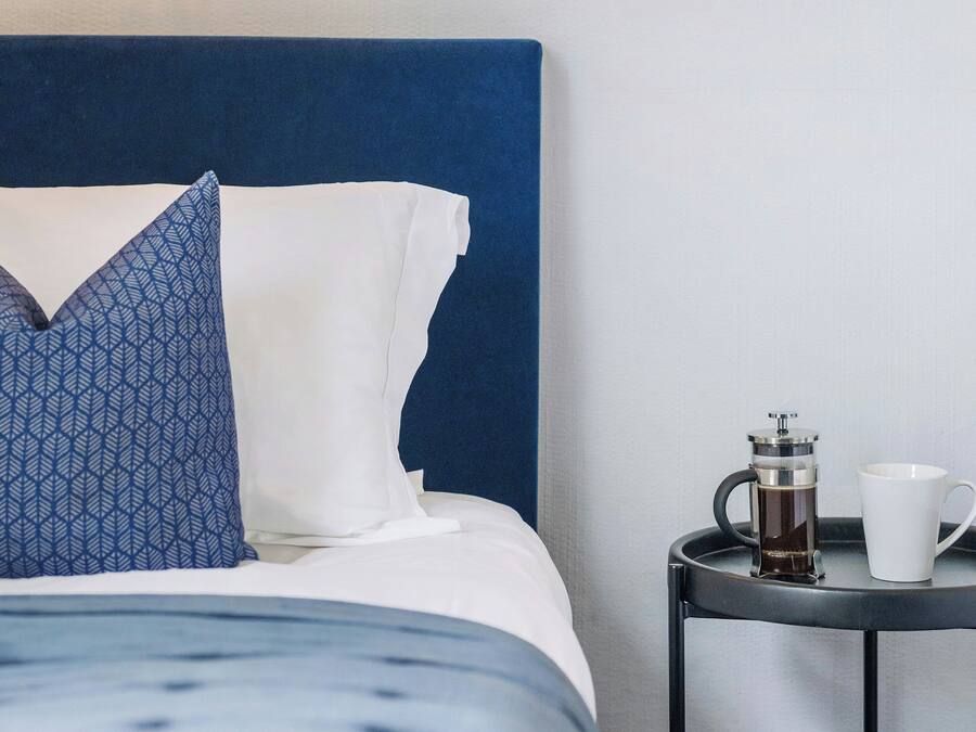 A neatly made bed with a blue headboard, a patterned pillow, and a white pillow. Beside the bed is a small table with a coffee maker and a white mug.