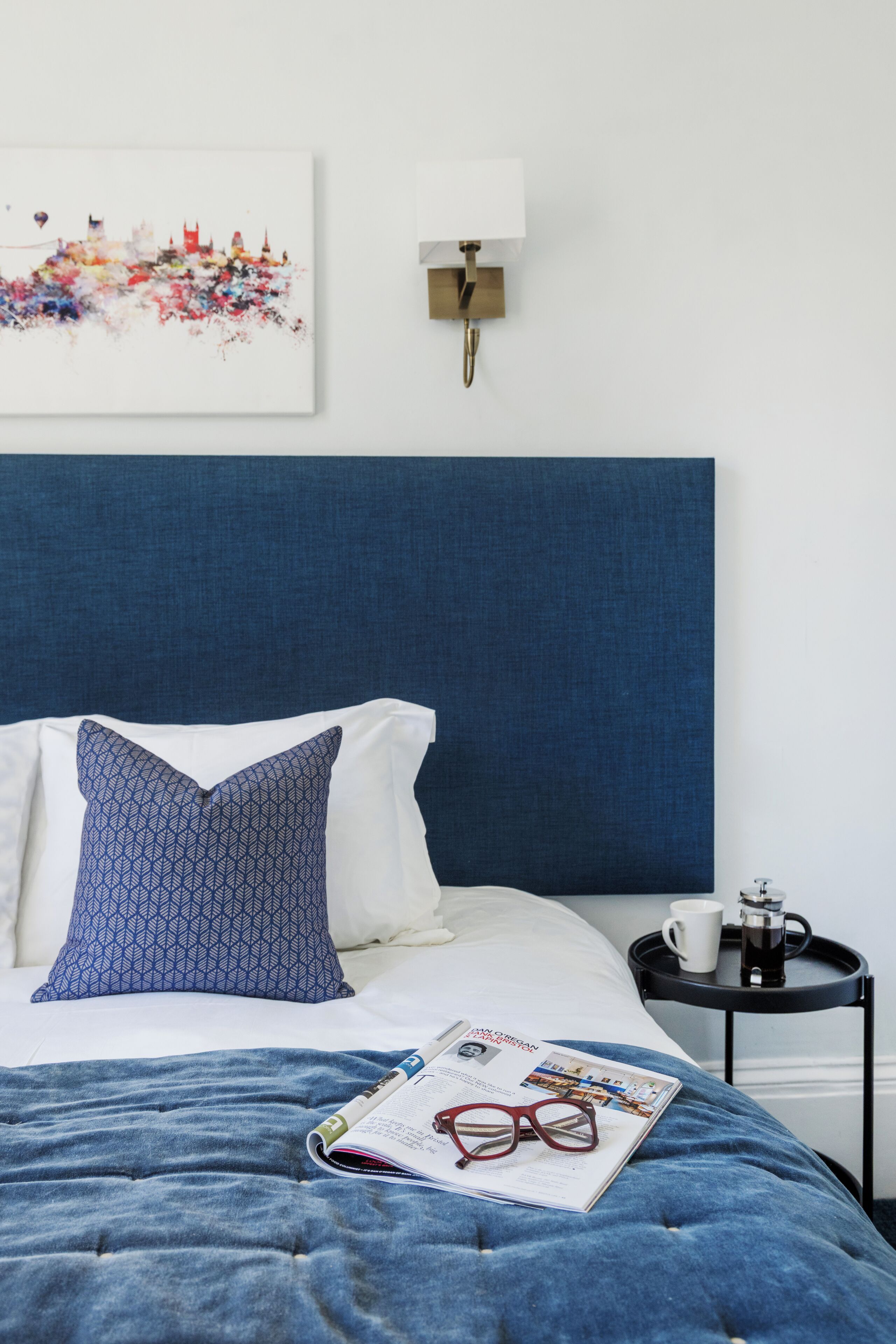 A neatly made bed with a blue and white patterned pillow, a magazine, and a pair of glasses on the bed. There is a small table with a cup and a coffee maker on it. A wall-mounted light fixture is visible above the bed.