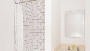 Un baño con inodoro blanco, ducha con cortina blanca, lavamanos con espejo y suelo de baldosas.