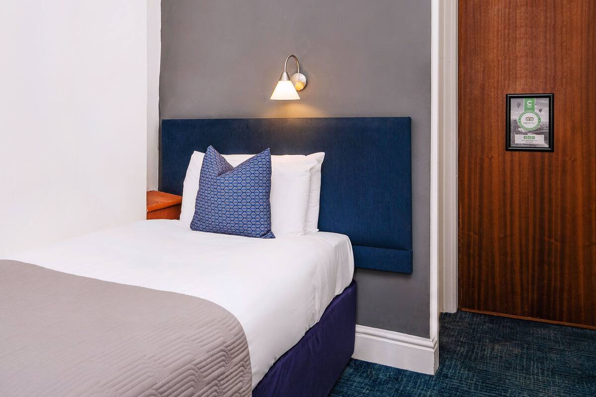 A hotel room with a bed, a nightstand, a wall-mounted lamp, and a framed certificate on the wall.