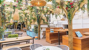 An outdoor seating area with wooden tables and chairs, surrounded by greenery and hanging plants.
