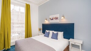Habitación de hotel con una cama grande, una mesita de noche, una lámpara de pared y un ventanal con cortinas amarillas.