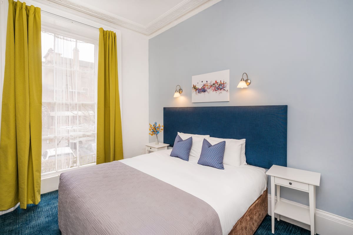 A hotel room with a large bed, a nightstand, a wall-mounted lamp, and a window with yellow curtains.