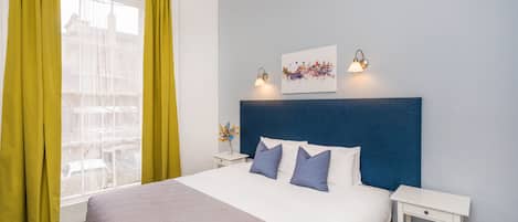 A hotel room with a large bed, a nightstand, a wall-mounted lamp, and a window with yellow curtains.