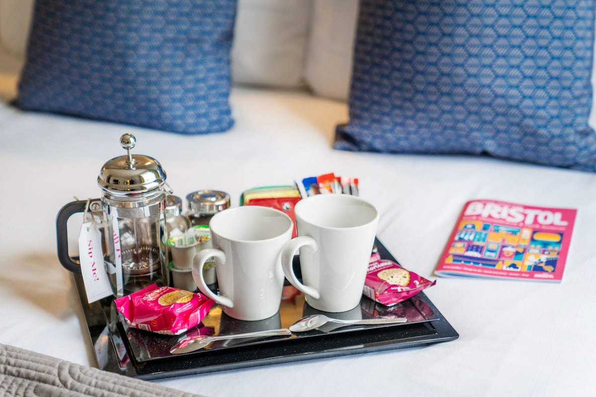 A tray with a coffee maker, two white mugs, a spoon, and some snacks on a bed with blue patterned pillows and a magazine titled "BRISTOL".