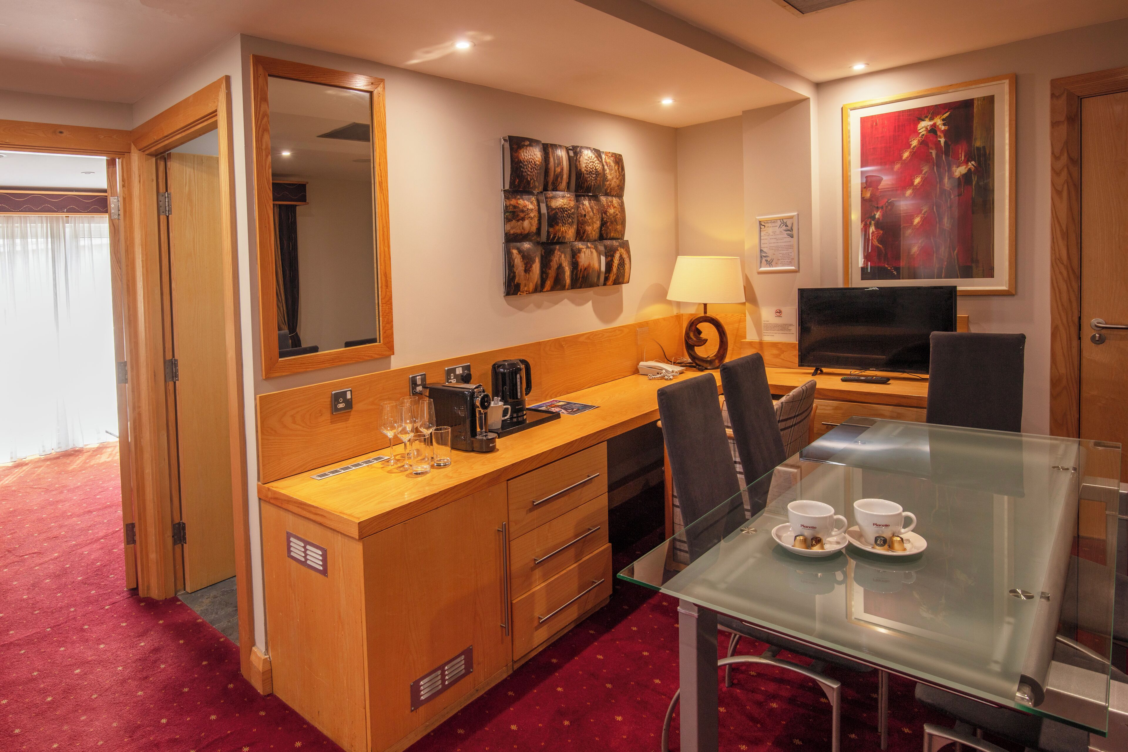 Habitación de hotel con un escritorio de madera, una mesa de vidrio con dos tazas, un televisor y un cuadro en la pared.