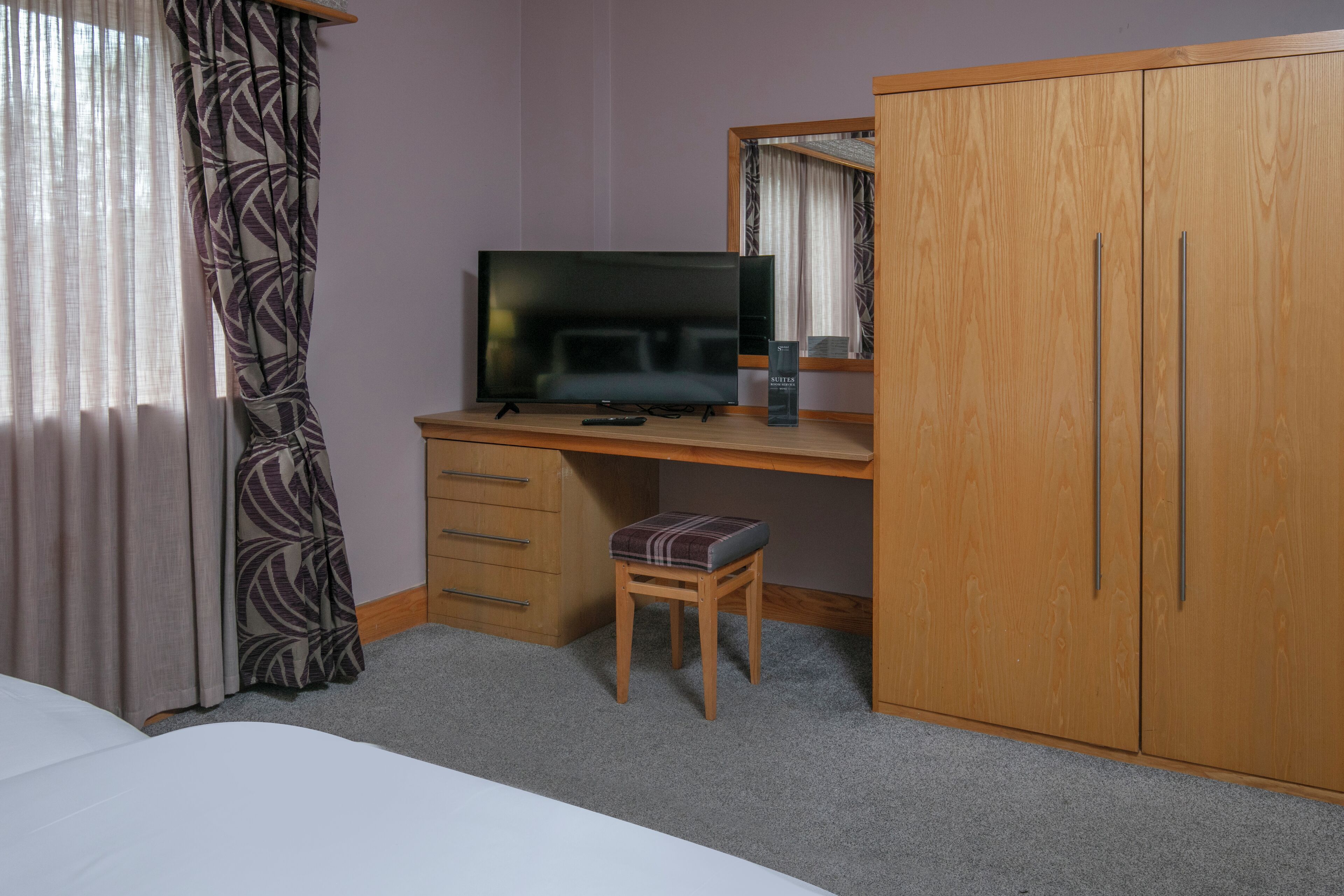 A hotel room with a wooden desk, a television, a small stool, a wardrobe, and a bed.