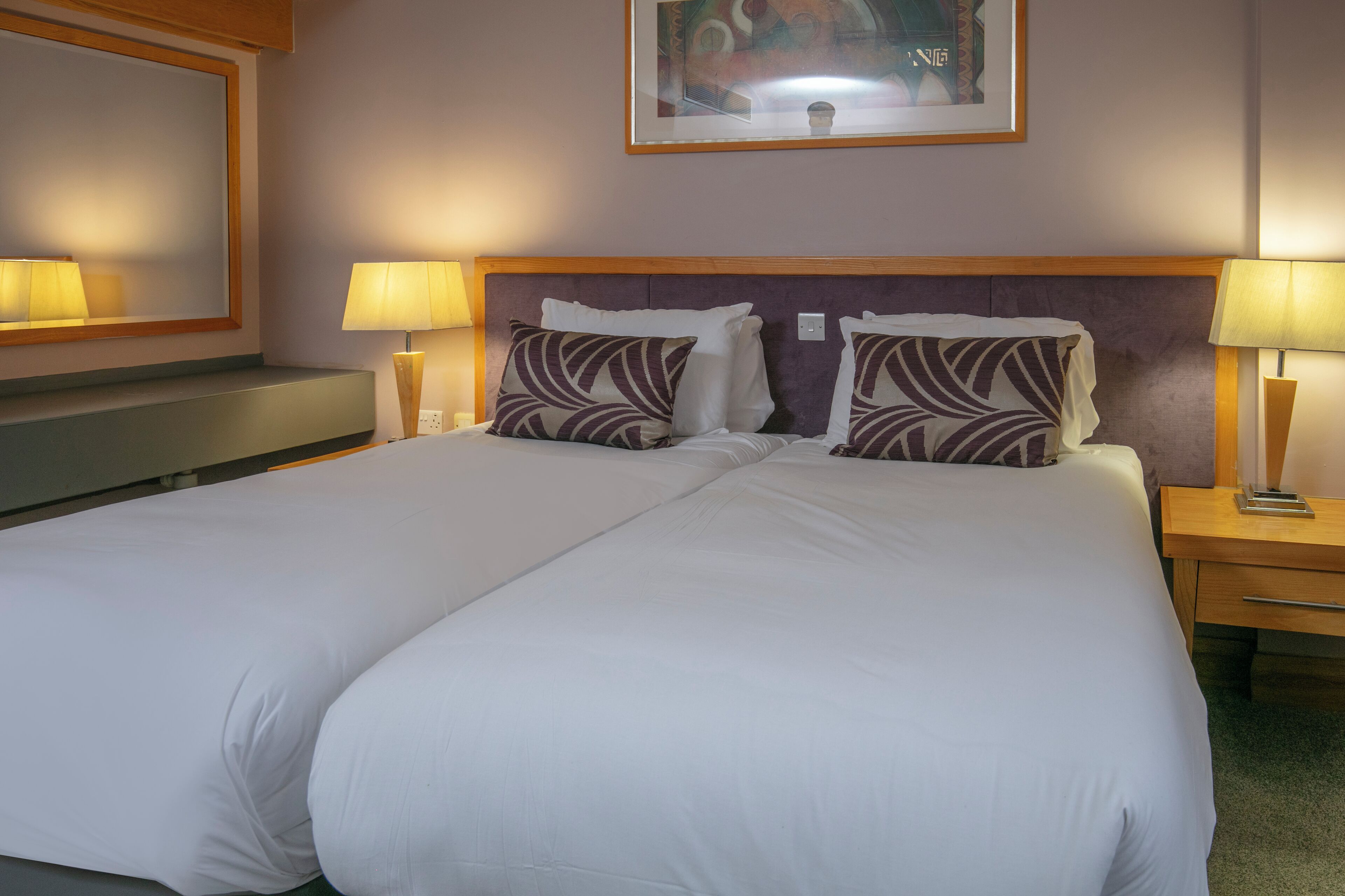 A double bed with white linens and patterned pillows, a nightstand with a lamp, and a painting on the wall.