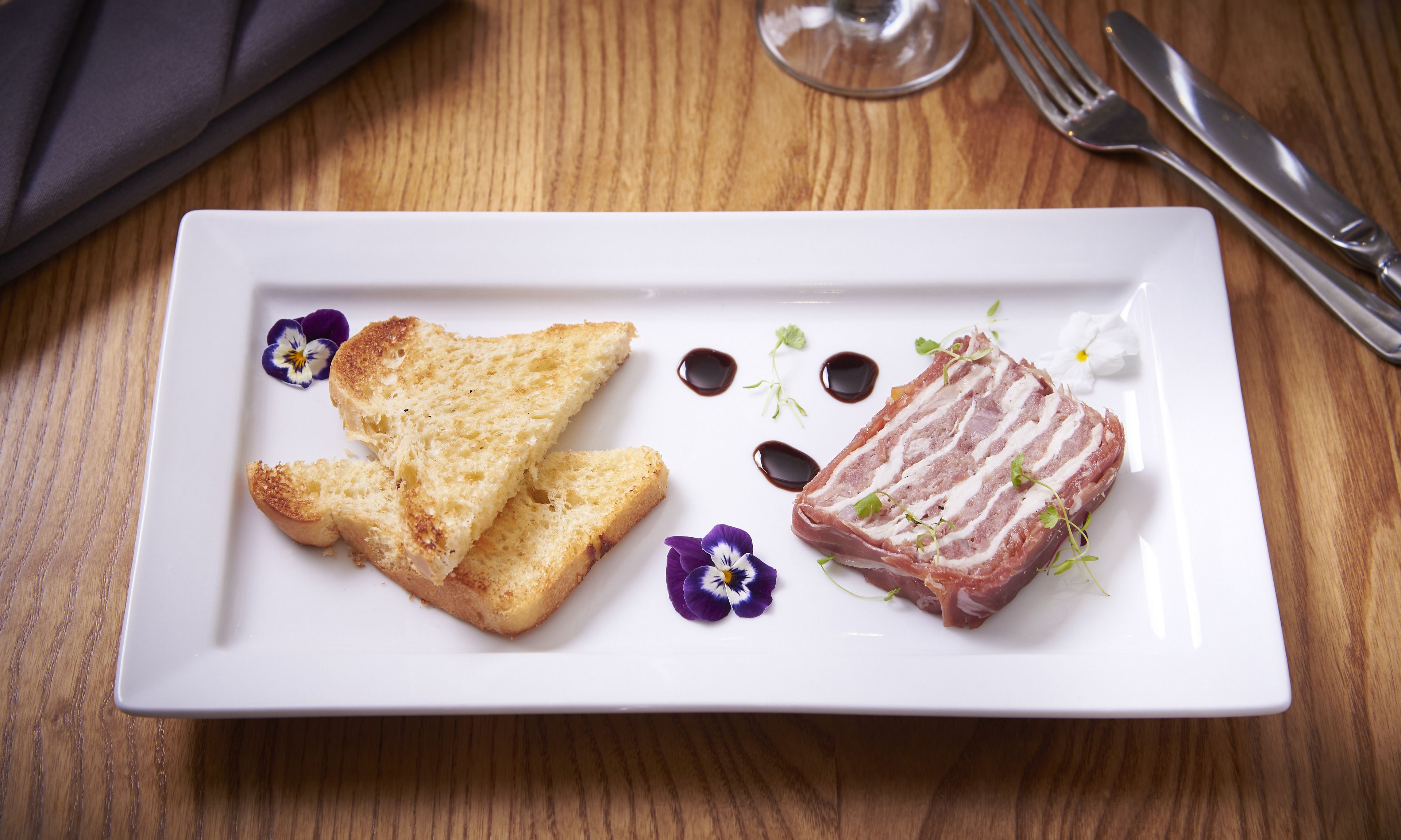 A white rectangular plate with toasted bread and a slice of meat, possibly bacon, on a wooden table.